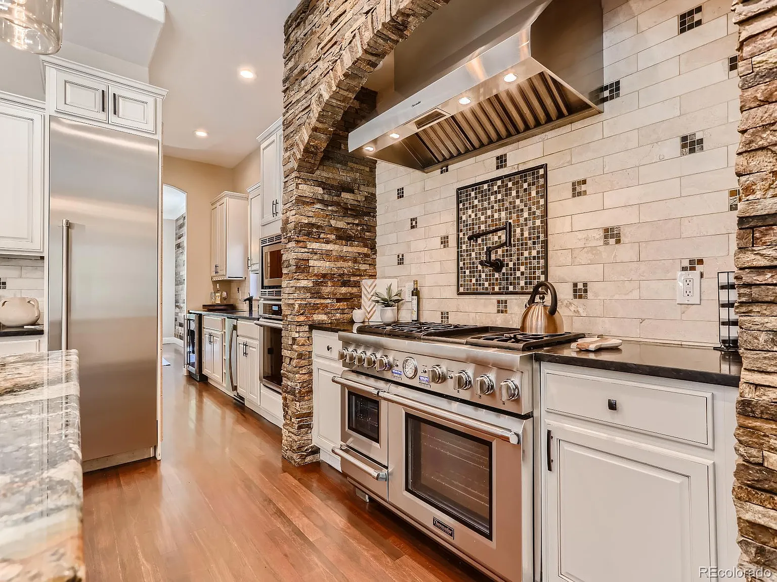 Luxury kitchen featuring a professional-grade stainless steel stove and hood, surrounded by a stone archway and tile backsplash with decorative accents, complemented by white cabinetry and hardwood floors.