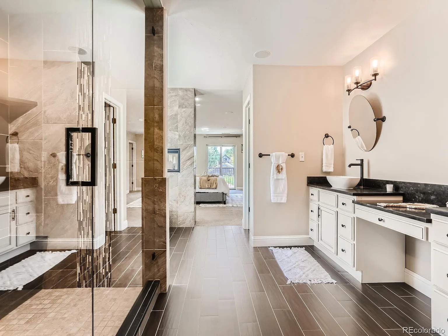 Luxurious bathroom featuring a spacious walk-in glass shower, double vanity with vessel sinks, circular mirrors, and sconce lighting, leading to a bedroom with a serene view.
