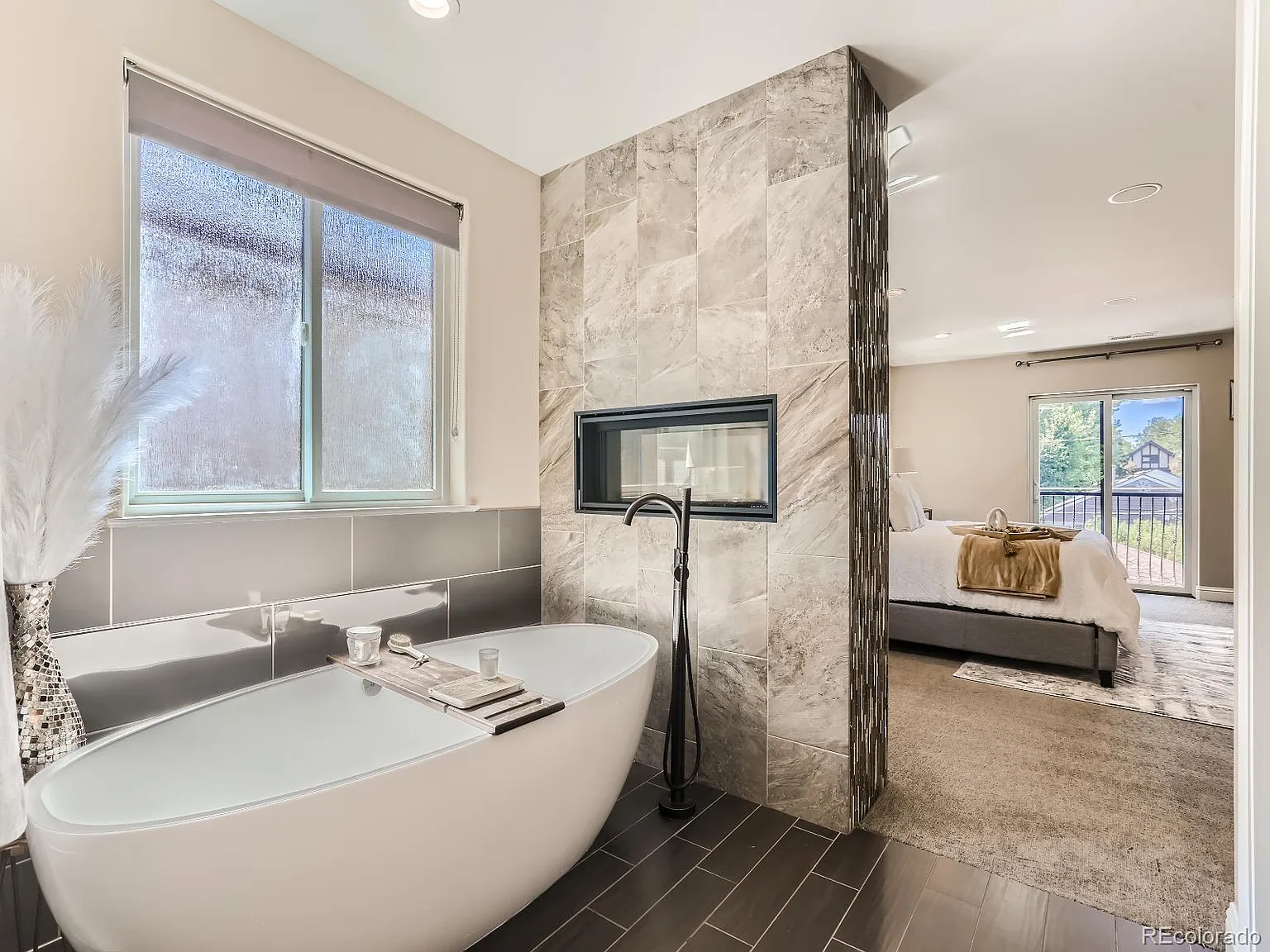 Modern bathroom with a freestanding oval bathtub, floor-mounted faucet, and a two-sided tiled fireplace divider, overlooking a connected bedroom with elegant decor.