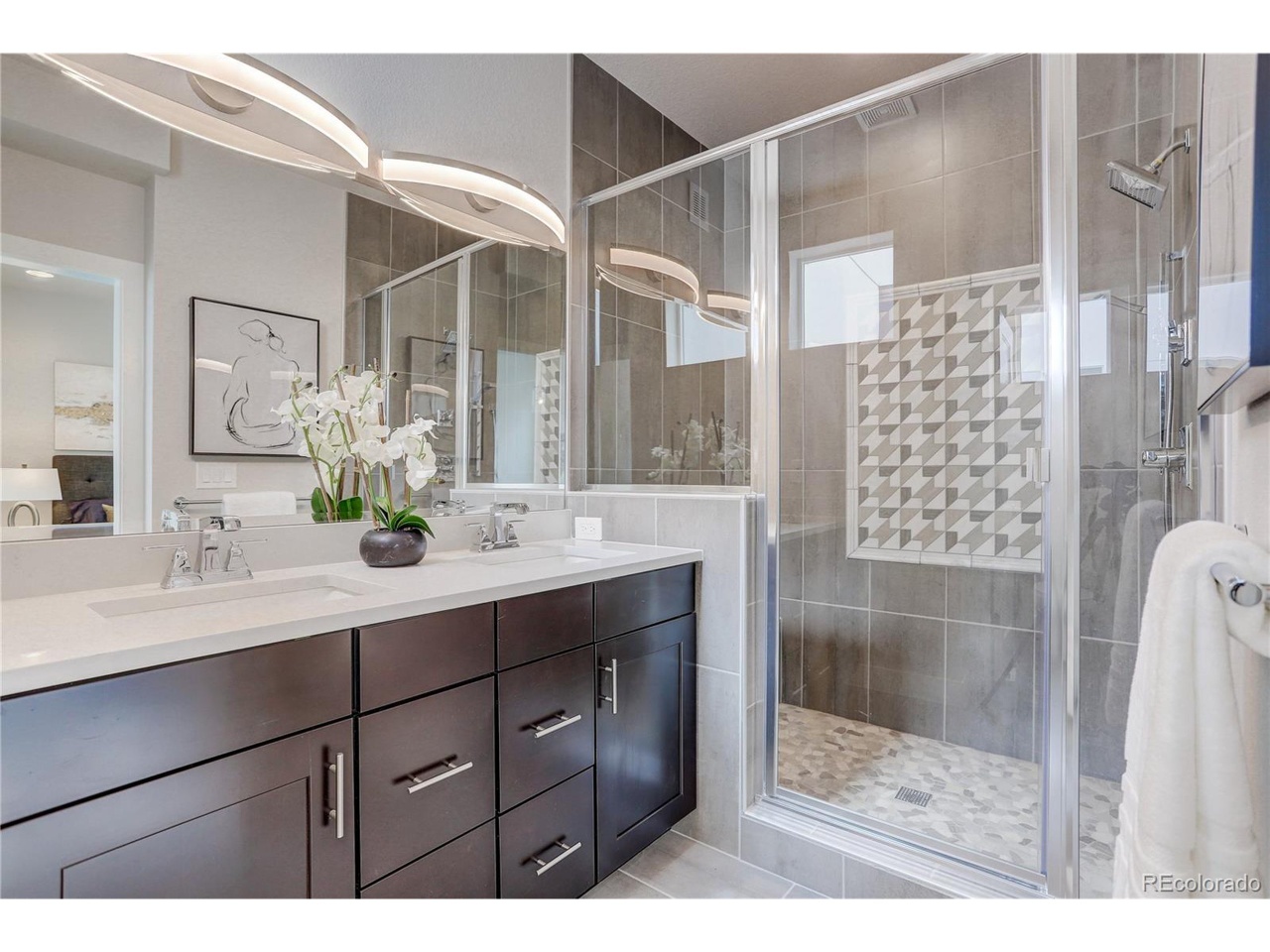 Modern bathroom with a dark wood double vanity, sleek white countertops, and a glass-enclosed shower featuring geometric tile accents.