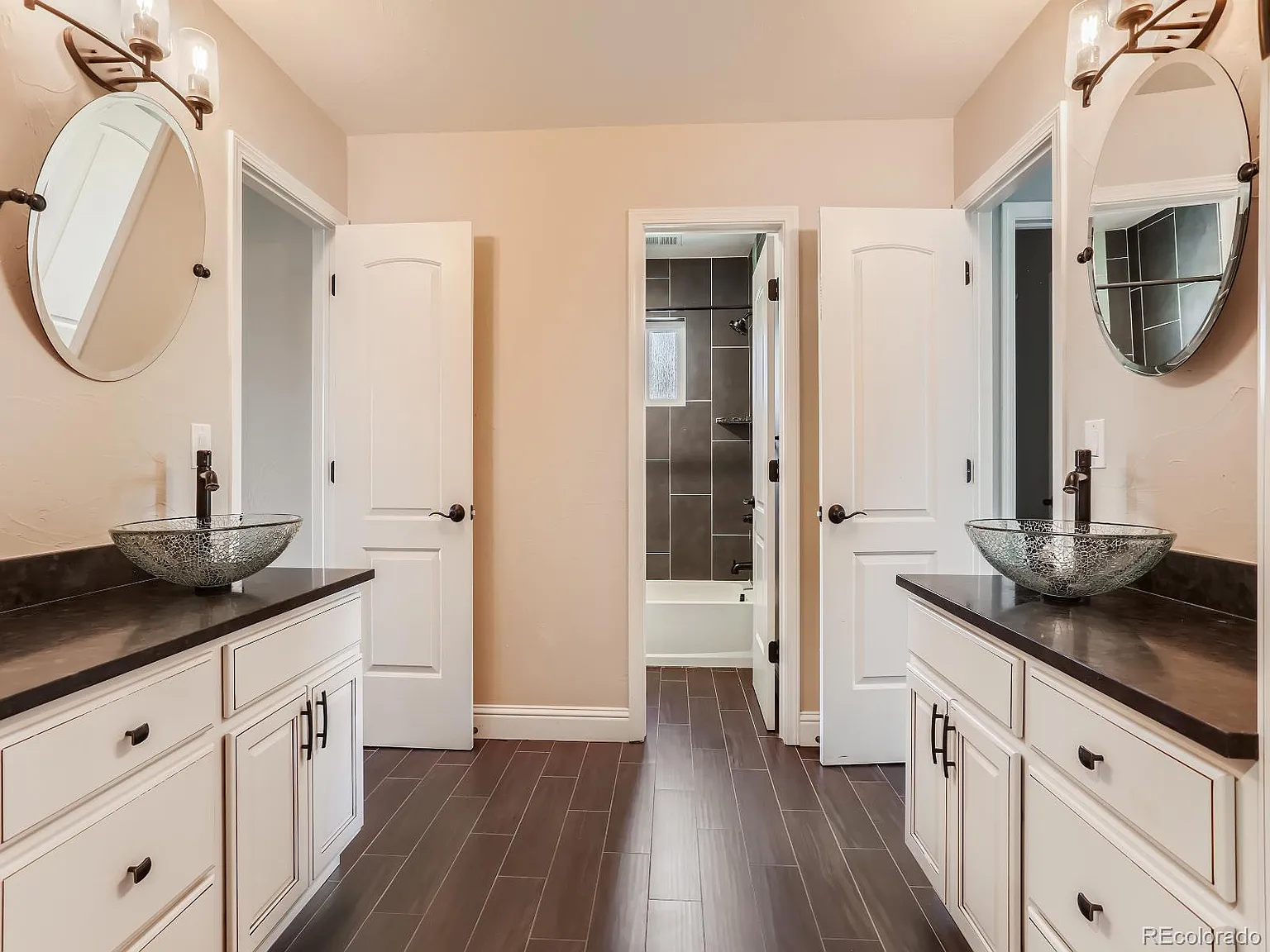 Elegant bathroom with dual white vanities, vessel sinks, round mirrors, and a doorway leading to a glass-enclosed shower area.