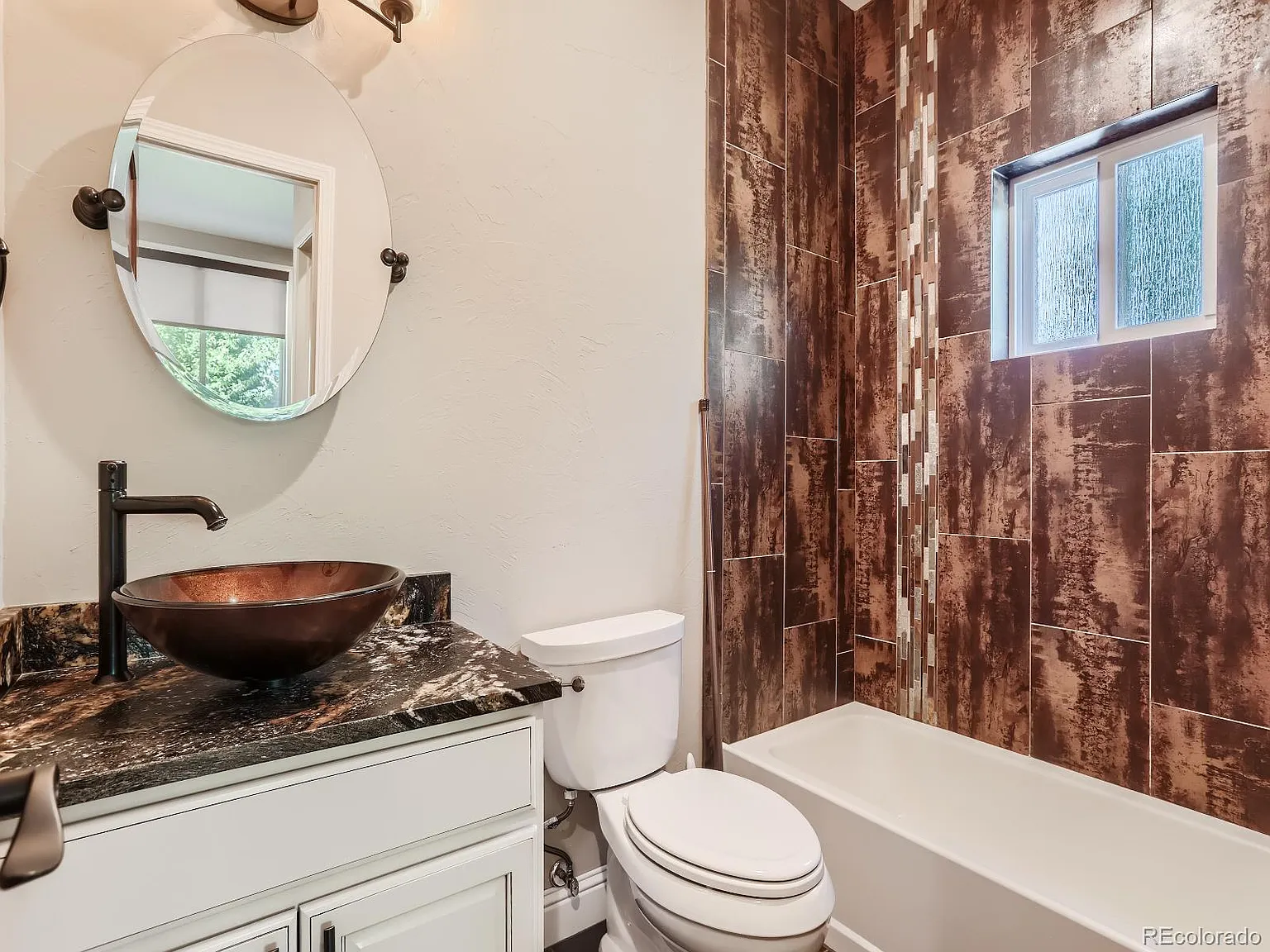 Stylish bathroom with a copper vessel sink on a granite countertop, a round mirror, and a tiled shower-bath combo featuring rich brown textures and a small frosted window.