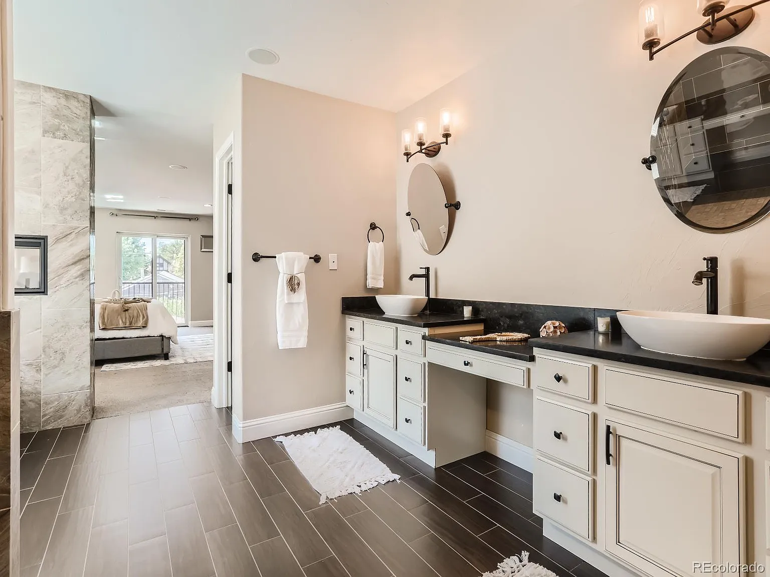 Elegant bathroom with dual vessel sinks on black granite countertops, round mirrors, and modern lighting. The space features wood-look tile flooring and an open layout leading to a luxurious bathtub and walk-in shower.