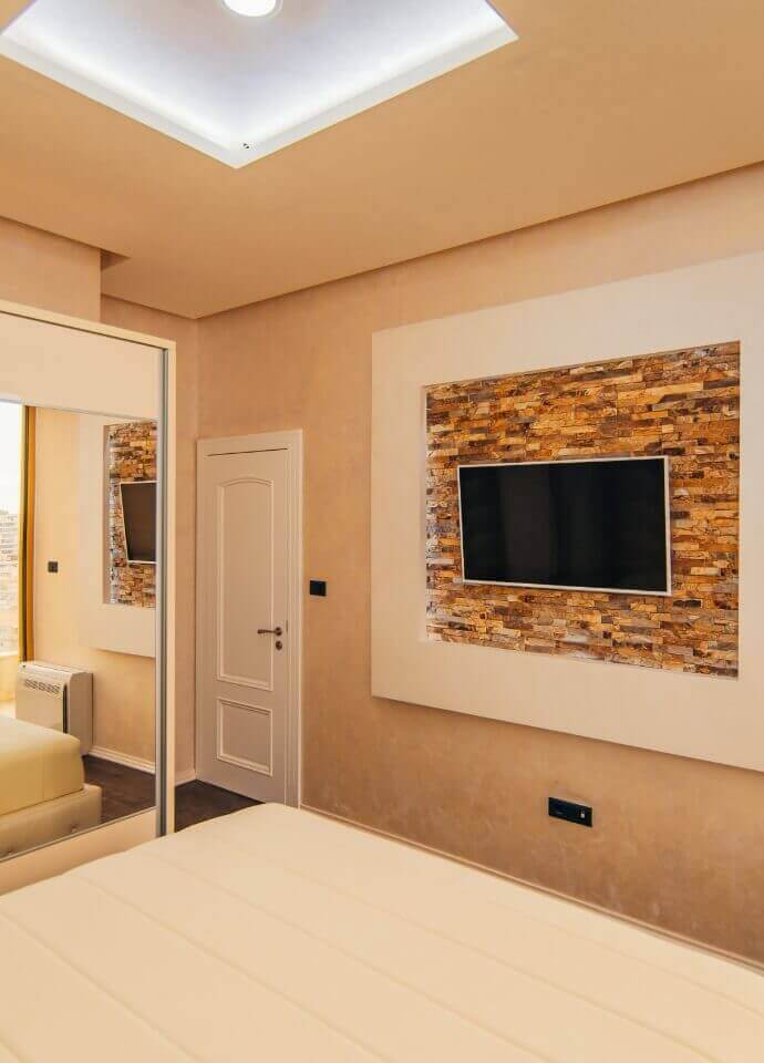 Bedroom with a wall-mounted TV set in a textured stone feature wall, contemporary ceiling lighting, and neutral decor tones.