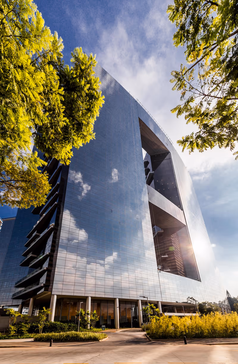 Modern glass skyscraper reflecting blue sky and clouds, surrounded by green trees and shrubs.