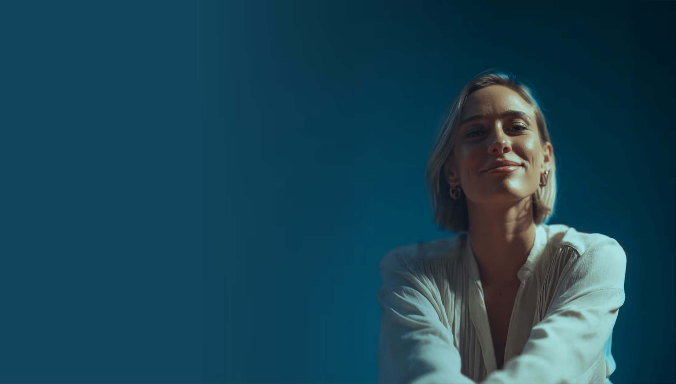 Mulher sorridente com cabelo loiro curto e blusa branca, sentada contra fundo azul escuro.