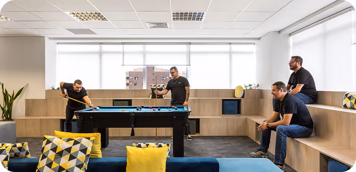 Quatro homens jogando bilhar em uma sala moderna com janelas grandes e almofadas coloridas.