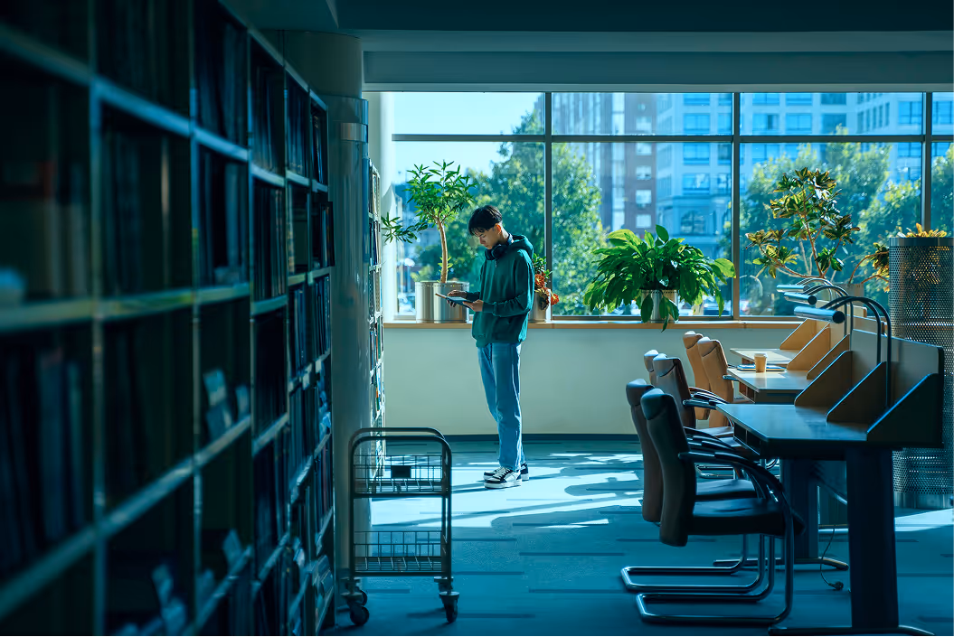 Jovem em camiseta verde está lendo um livro enquanto está em uma biblioteca com grandes janelas e plantas ao fundo.