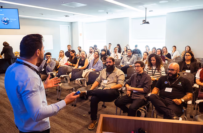 Homem falando para público atento em sala clara com cadeiras em estilo auditório.