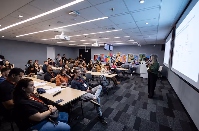 Palestrante apresentando slides para uma plateia atenta em uma sala de conferências moderna com luzes de LED no teto.