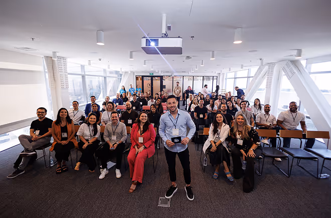 Grupo diversificado de pessoas sentadas em uma sala de conferências moderna, com um homem sorridente em pé na frente segurando um crachá.