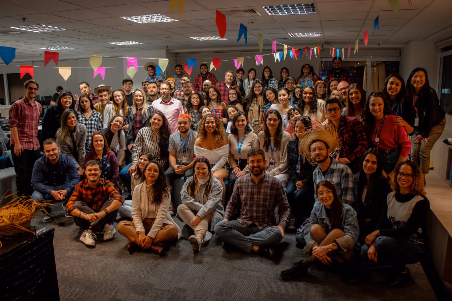 Grupo grande de pessoas reunidas em ambiente interno decorado com bandeirinhas coloridas, celebrando uma festa junina.