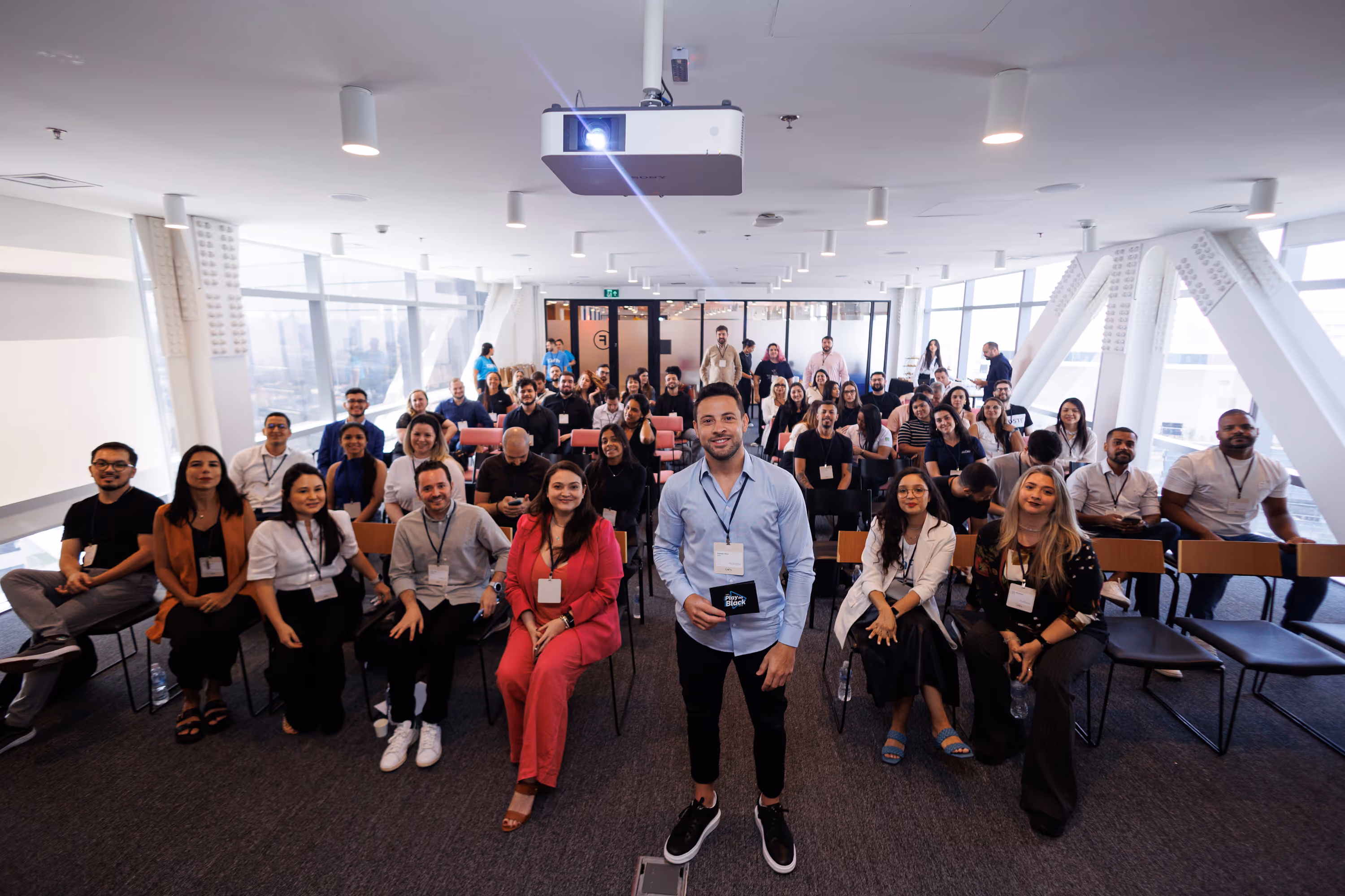 Grupo diverso de pessoas sentadas em cadeiras dentro de uma sala de conferências, com um homem em camisa azul clara em pé à frente segurando um cartão.