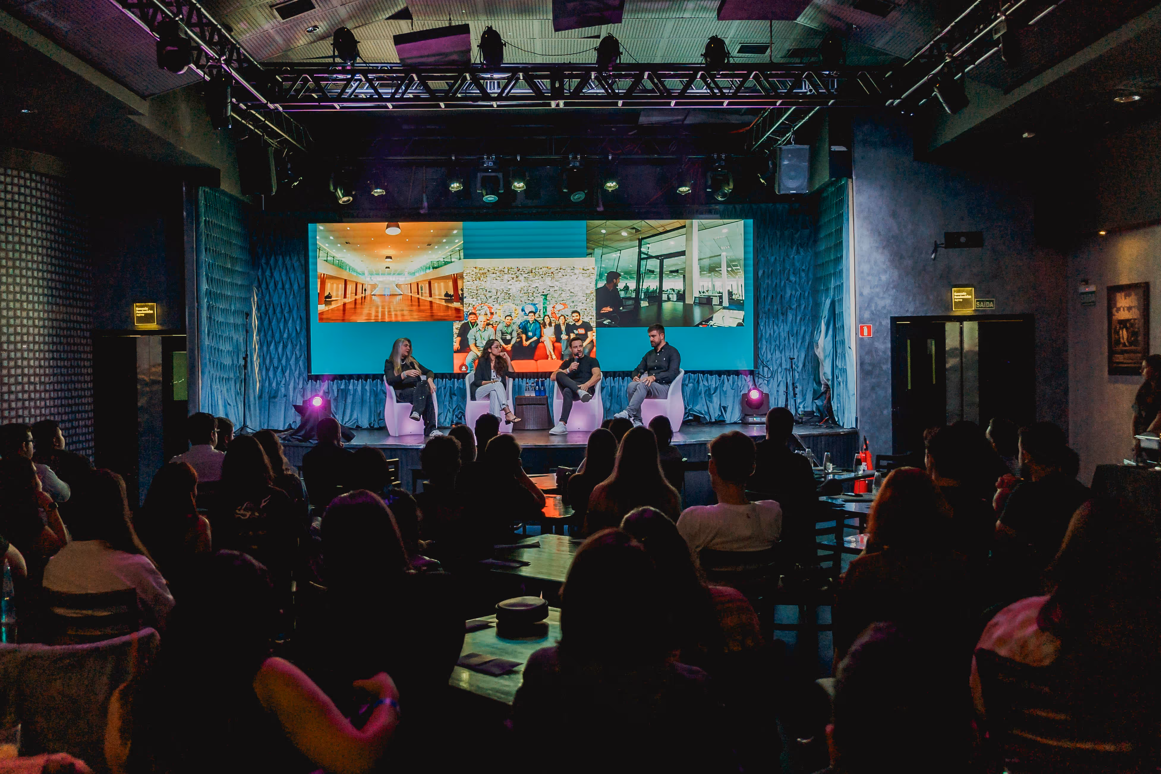 Quatro palestrantes sentados em cadeiras brancas no palco discutindo durante evento com tela grande ao fundo e público sentado em um ambiente escuro.