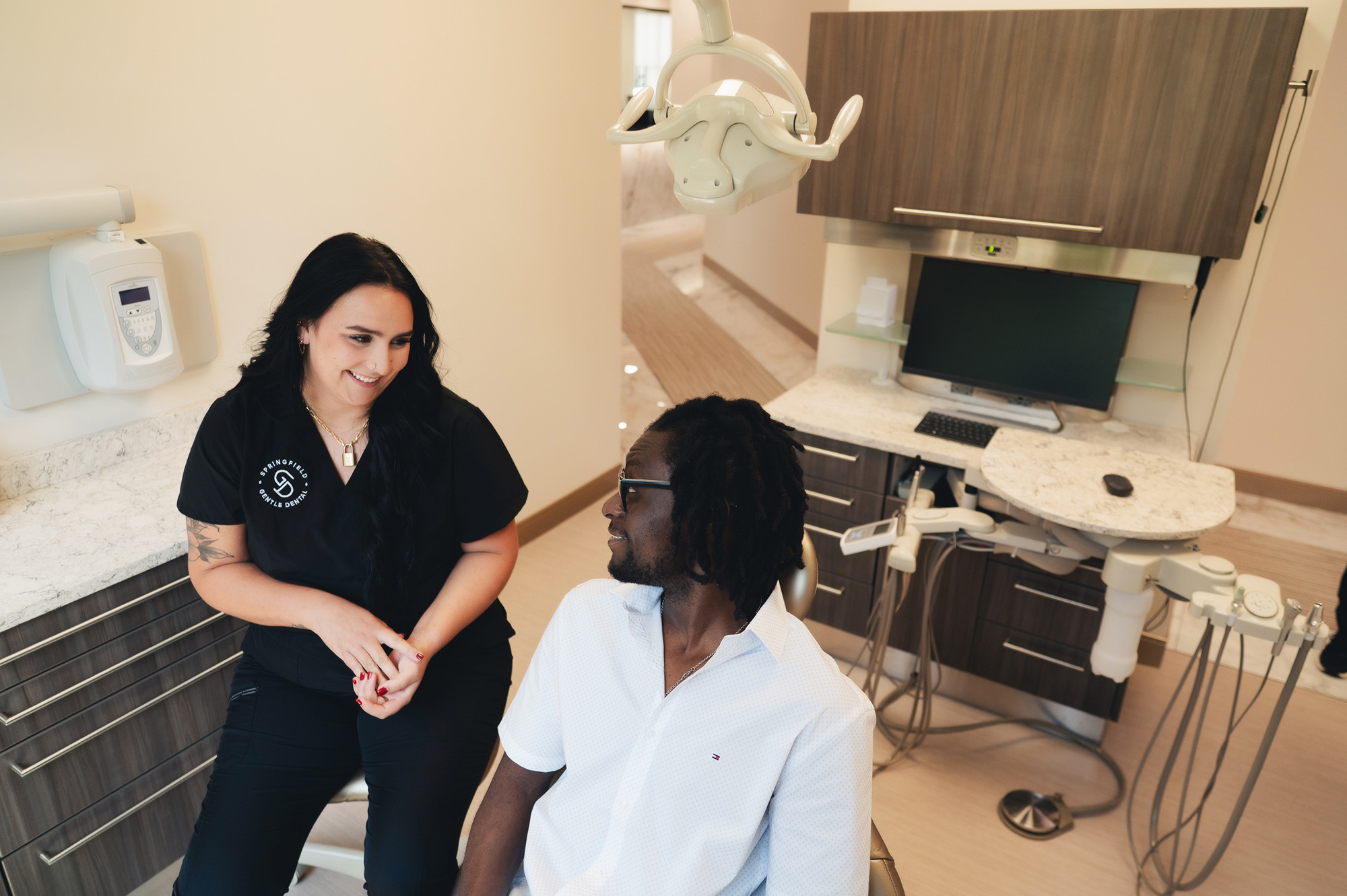 Dental professional in black scrubs smiling and talking with a male patient seated in a dental chair in a modern clinic.