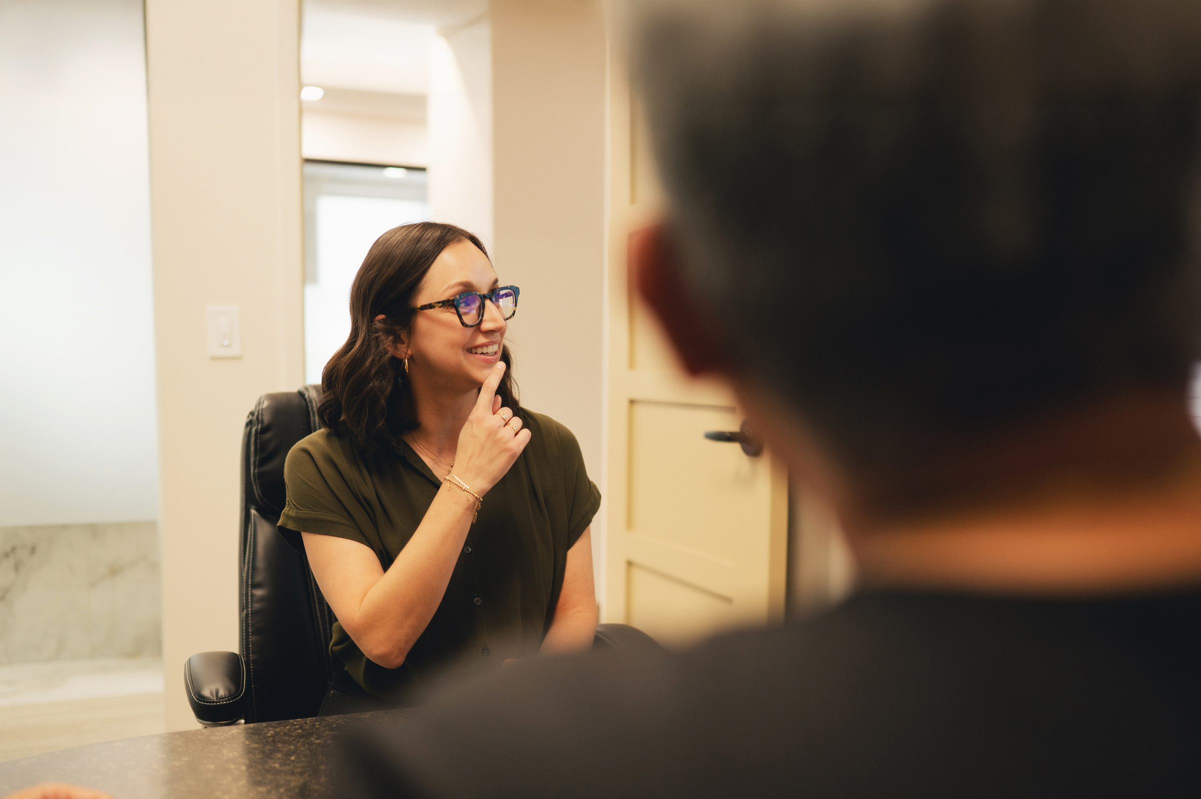 Smiling woman with glasses touching her chin, sitting in an office chair during a conversation.