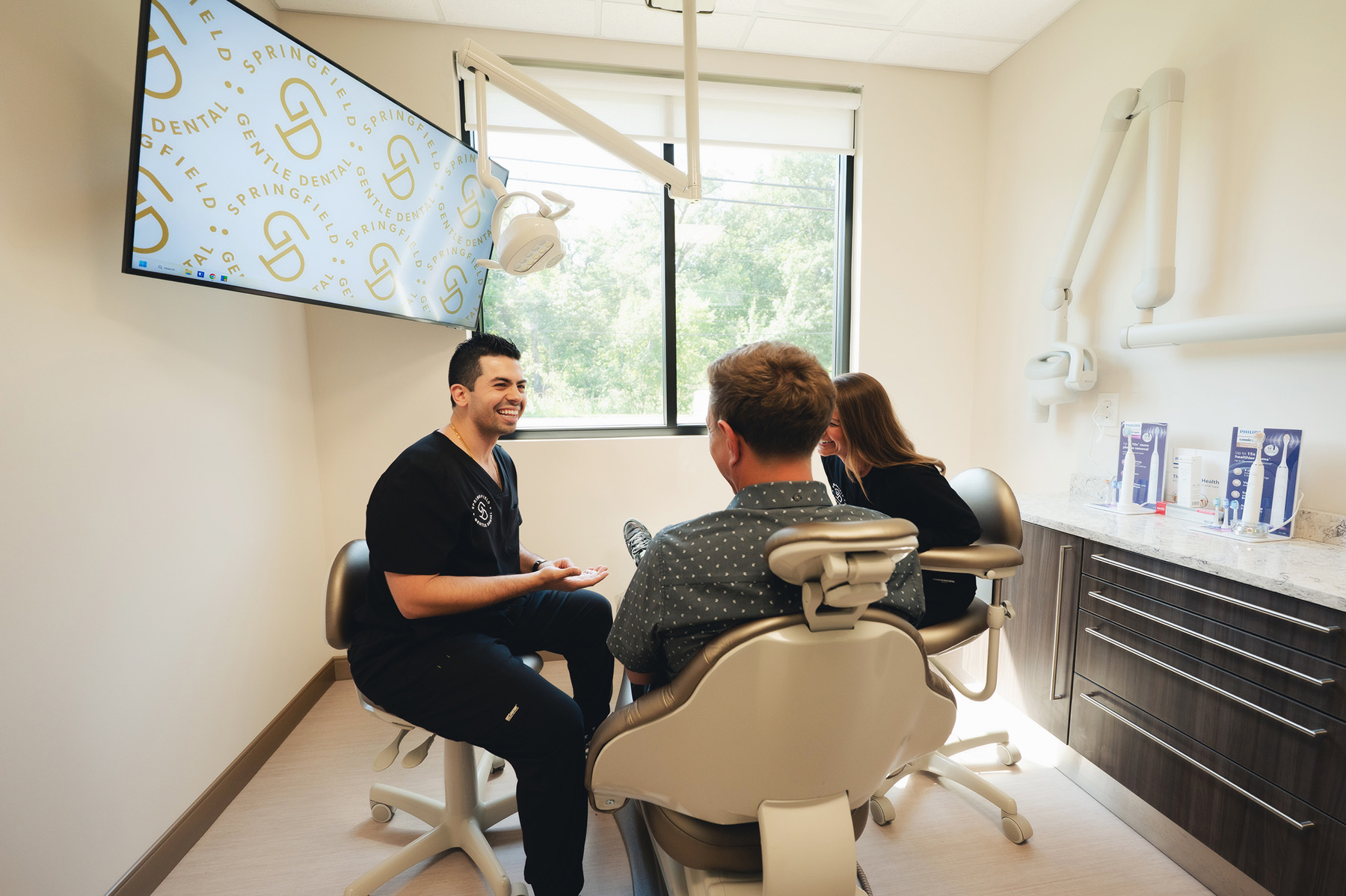 Dentist and assistant smiling and talking with a male patient seated in a dental chair in a bright clinic room.