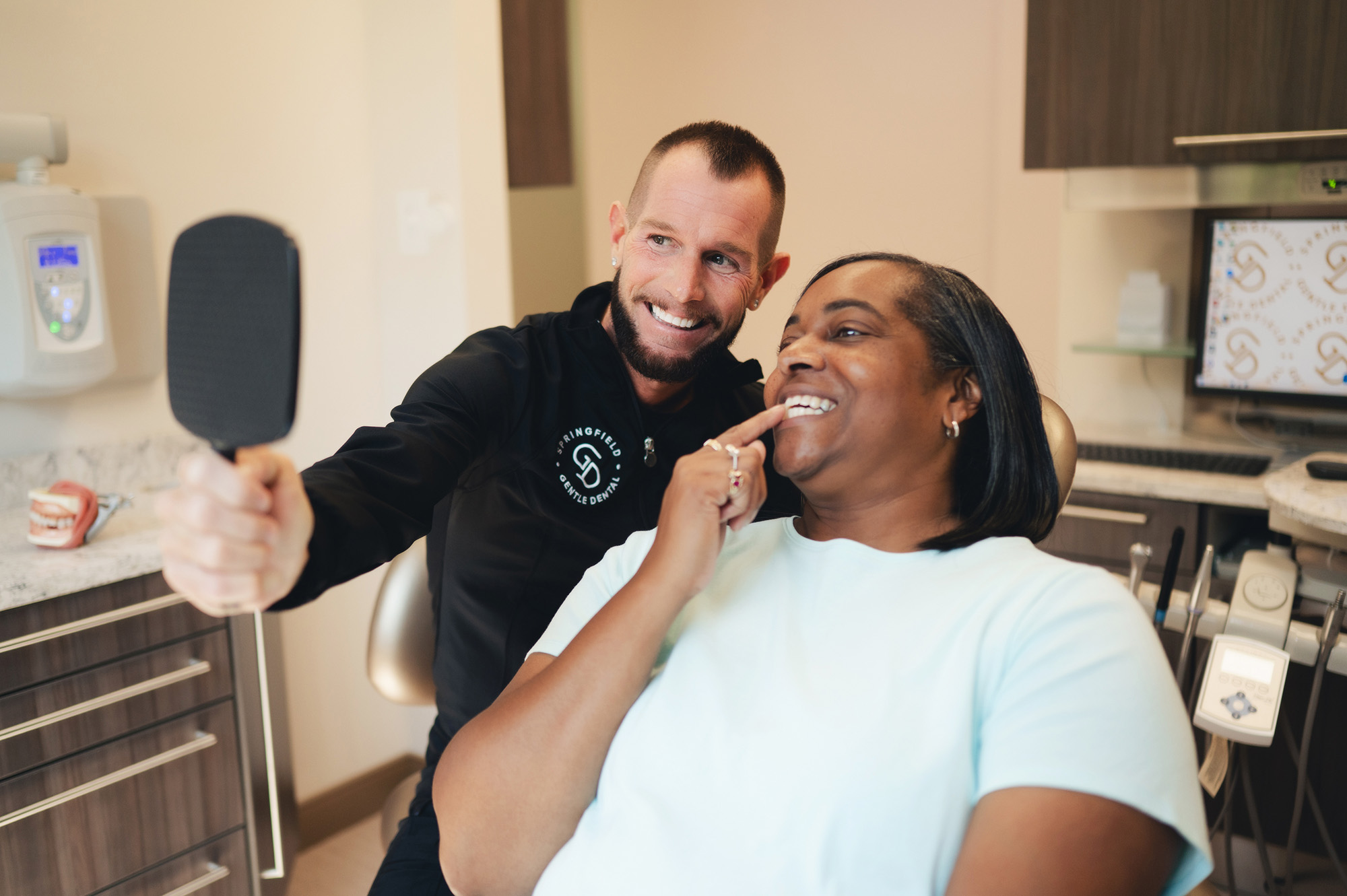 Dentist showing a smiling female patient her teeth in a hand mirror inside a dental office.