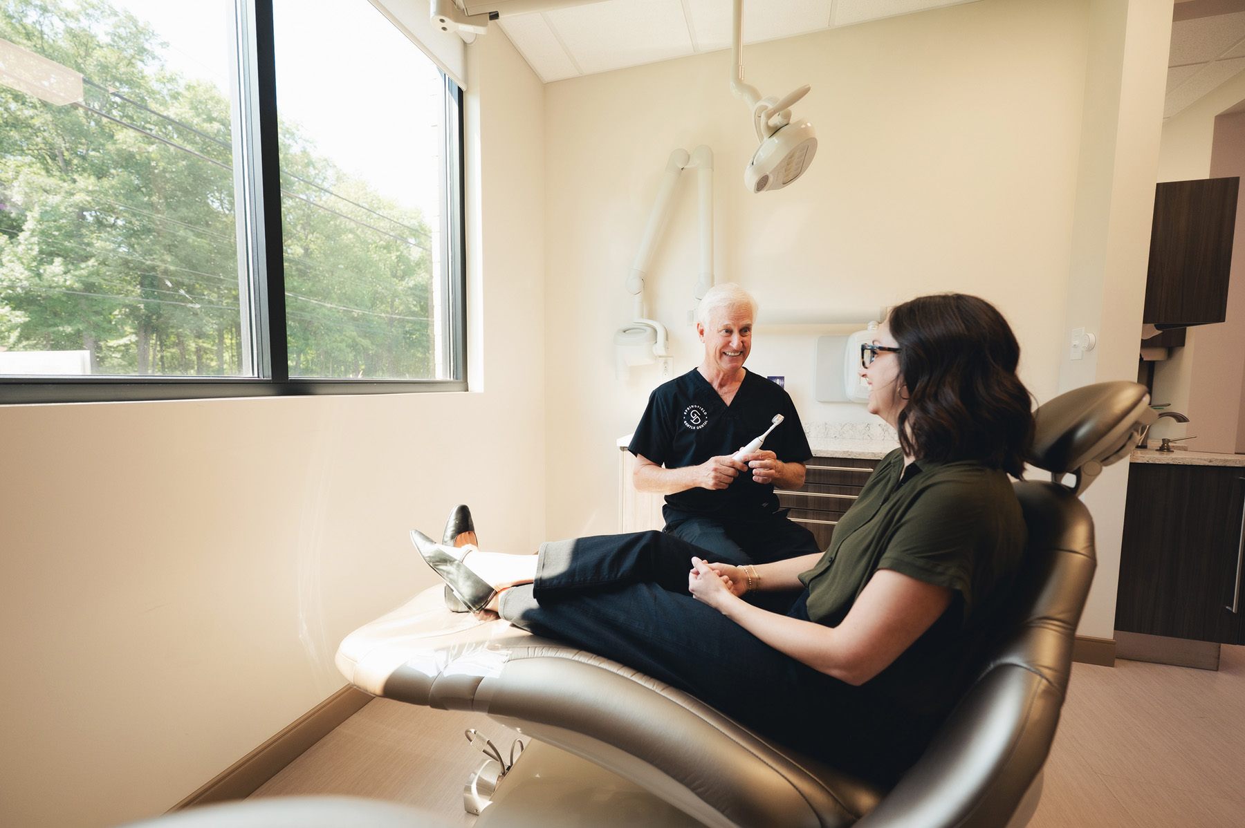 Dentist explaining an electric toothbrush to a female patient seated in a dental chair in a bright office.