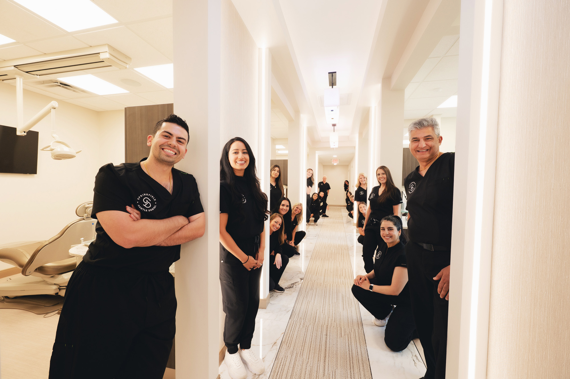 Dental clinic team in black scrubs smiling and posing in a bright hallway with treatment rooms on both sides.