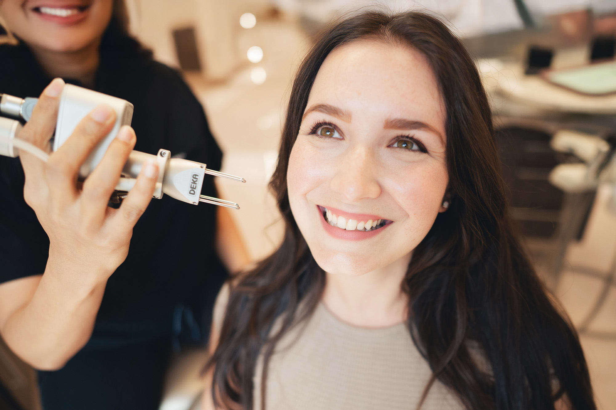 Smiling woman with long dark hair receiving a skin treatment using a DEKA device held by a professional.