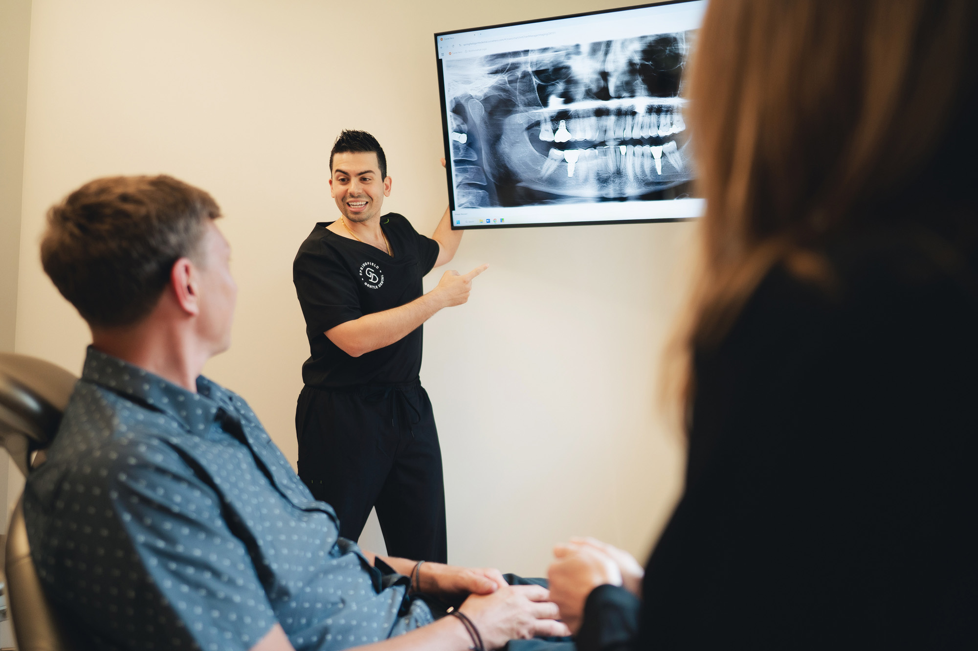 Dentist in black scrubs explaining a dental X-ray on screen to a seated patient and companion.