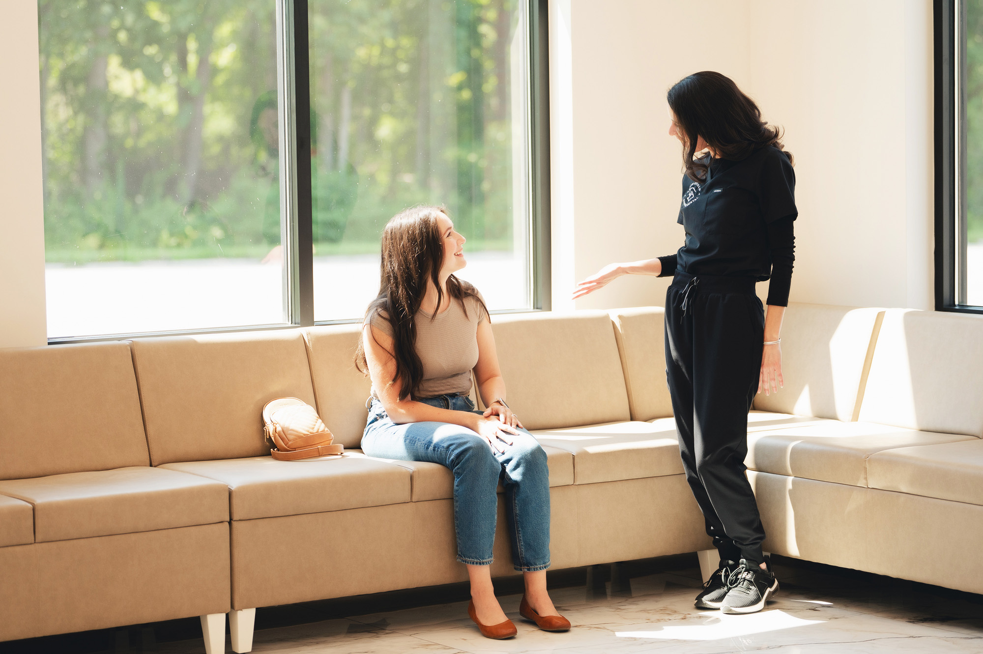 Two women talking in a sunny room with beige sofas and large windows overlooking greenery.