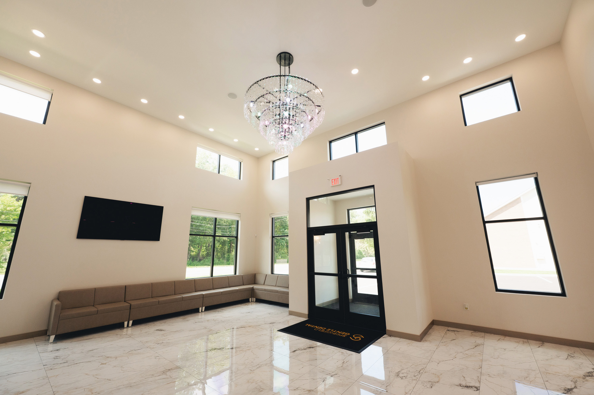 Modern waiting area with beige sectional sofa, large windows, marble floor, and a crystal chandelier.