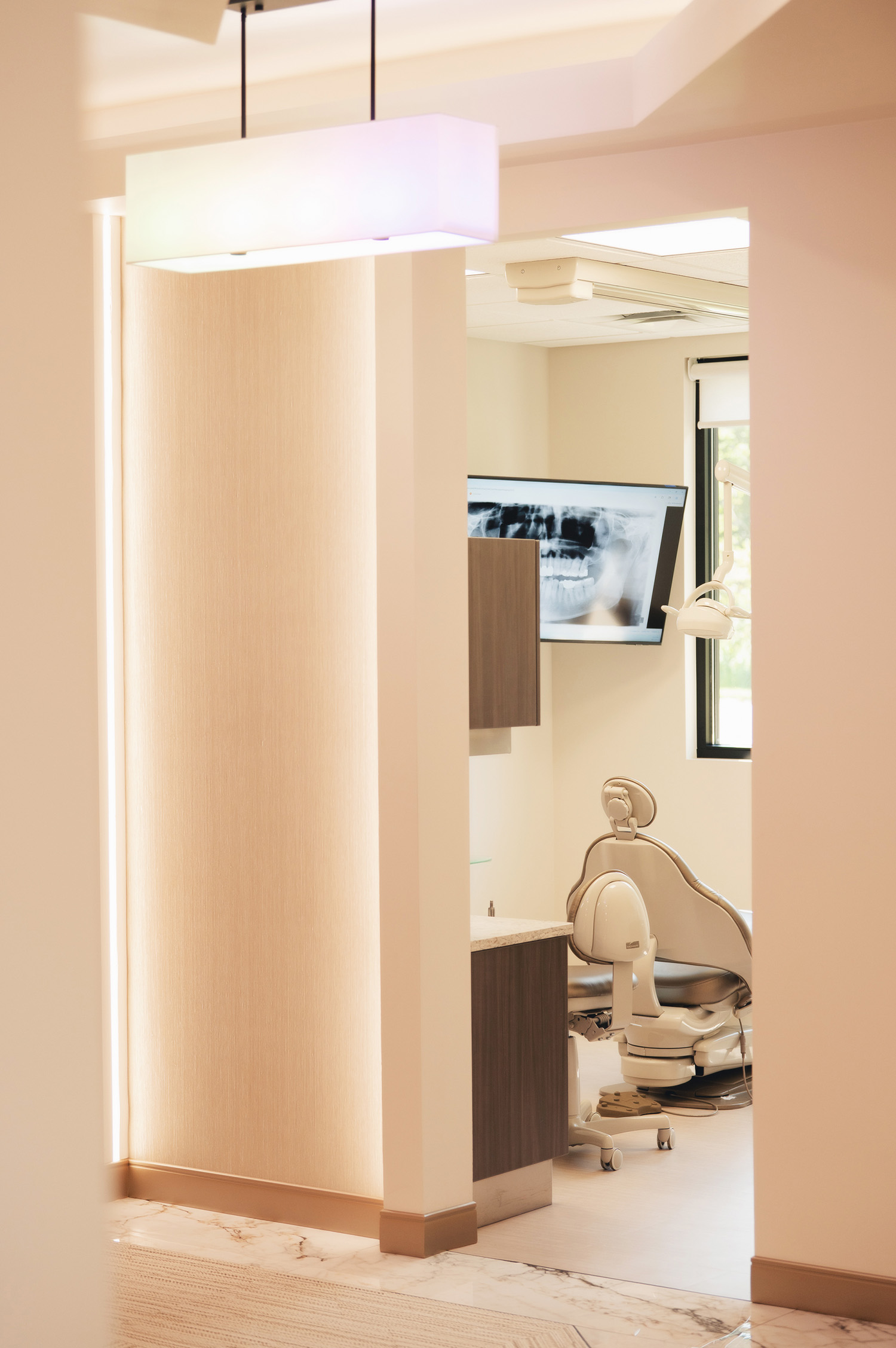View into a dental examination room featuring a dental chair, equipment, and a digital screen displaying a dental X-ray.