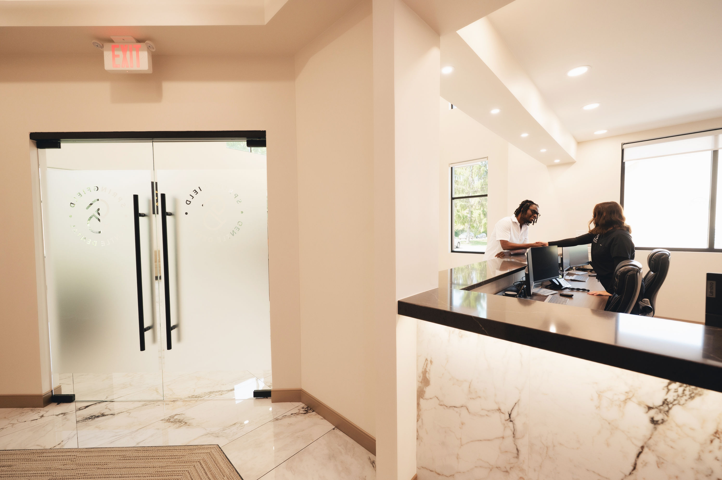 Reception area with marble desk where a staff member greets a smiling visitor across the counter in a bright office.
