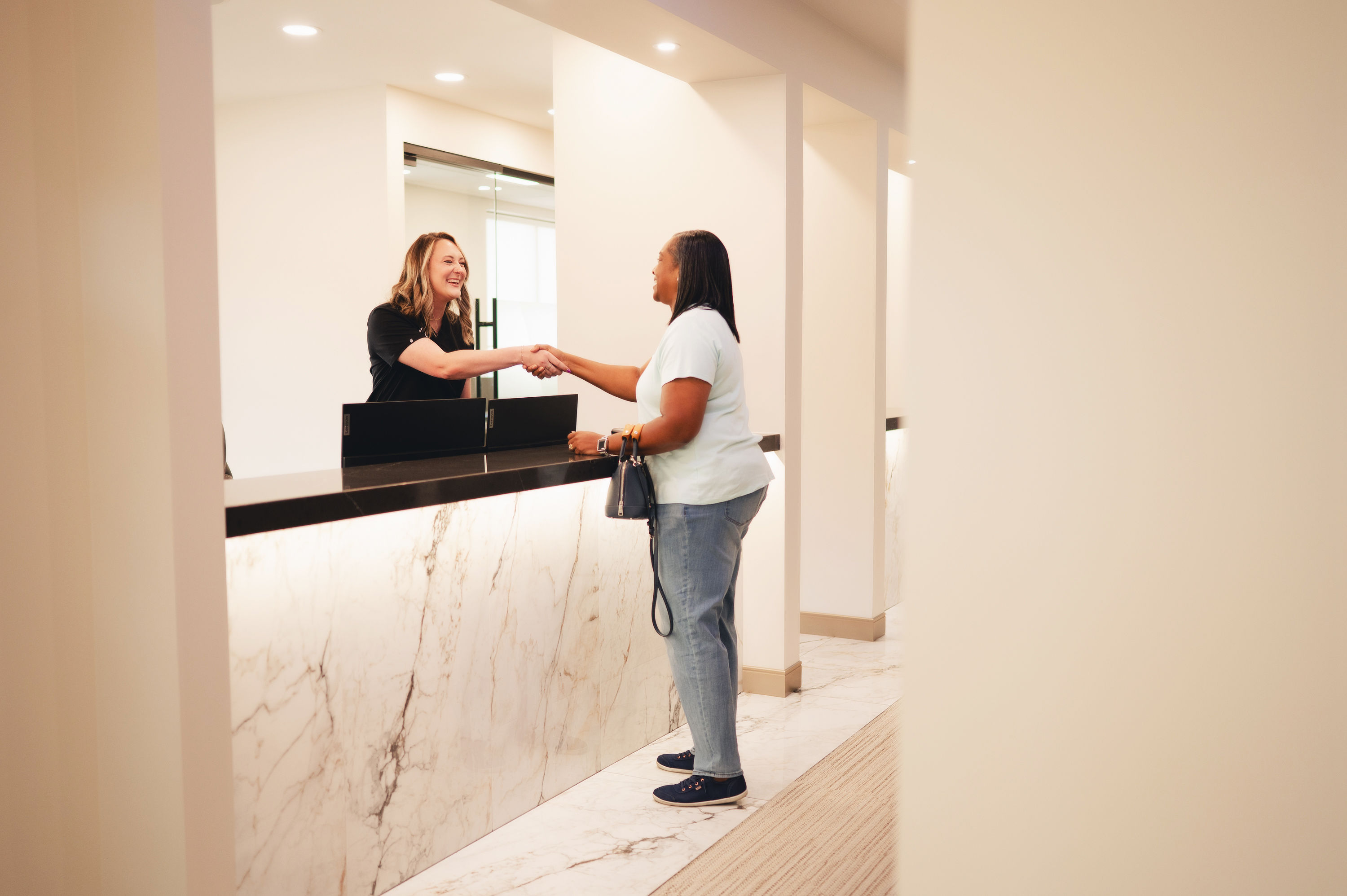 Receptionist and patient smiling and shaking hands across a marble reception desk in a bright office.