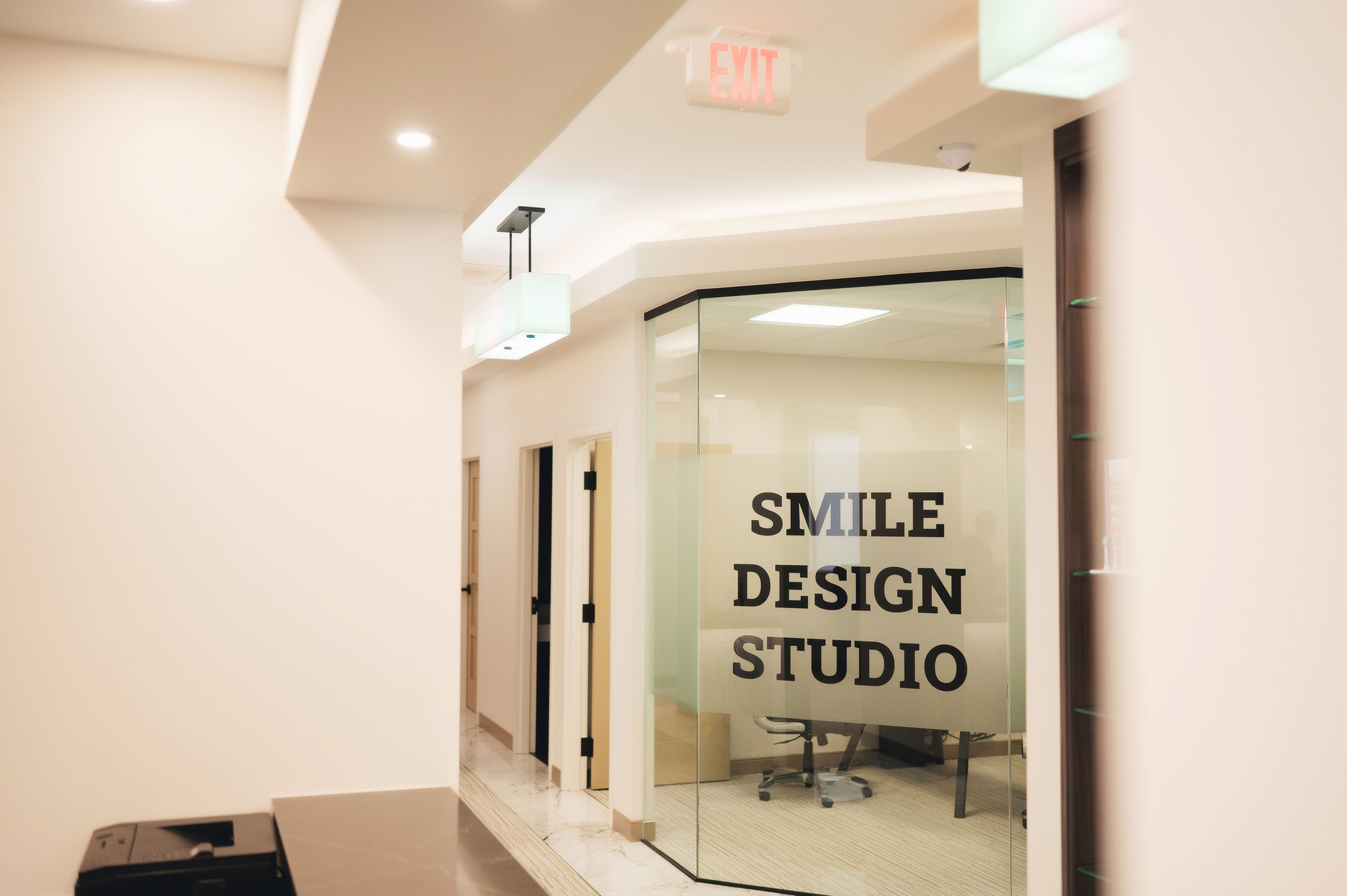 Modern clinic hallway with glass office labeled Smile Design Studio and an illuminated exit sign above.
