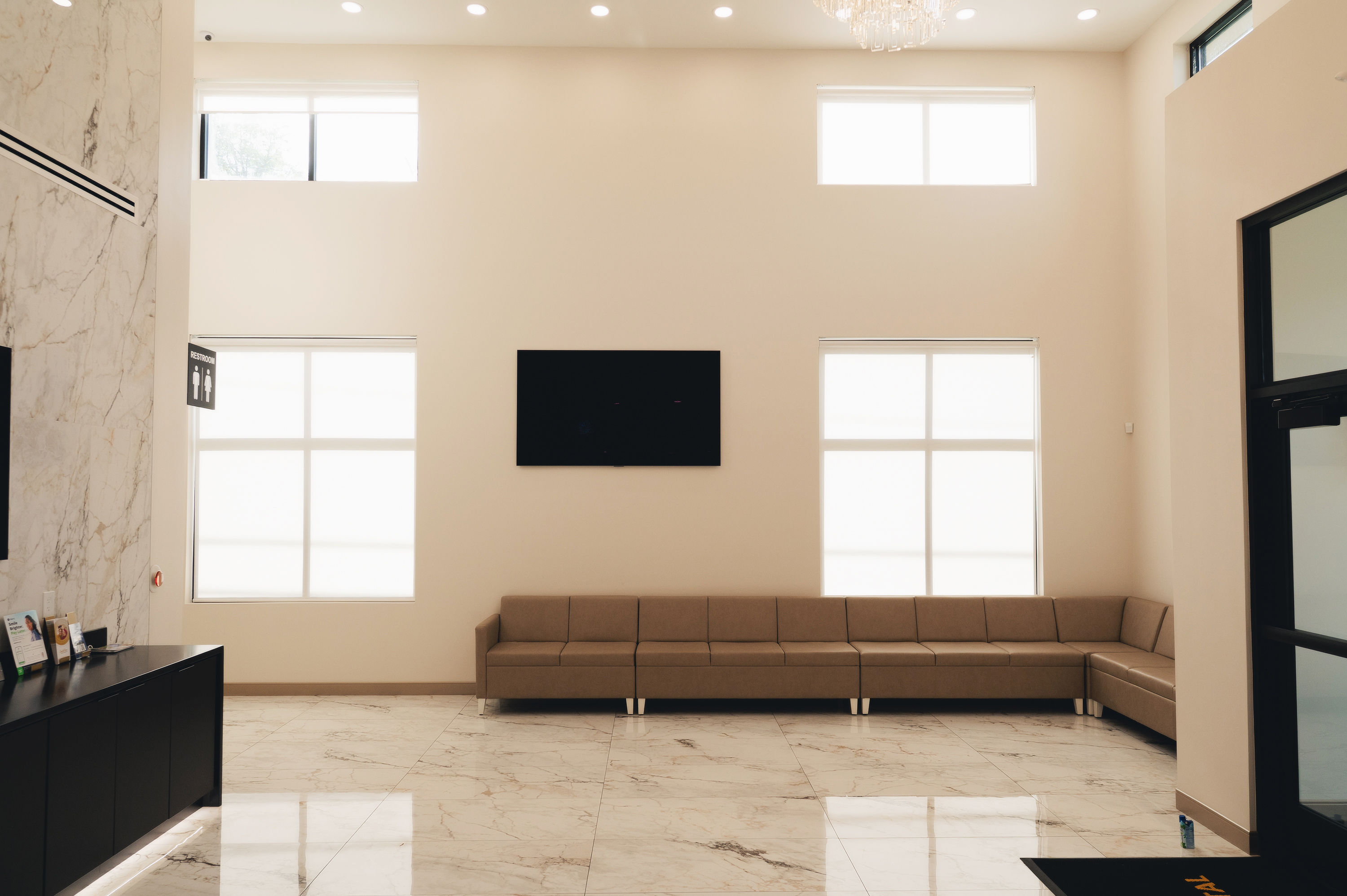 Modern waiting room with a long brown sectional sofa, marble floor, two large windows, a black wall-mounted TV, and a restroom sign on the left wall.