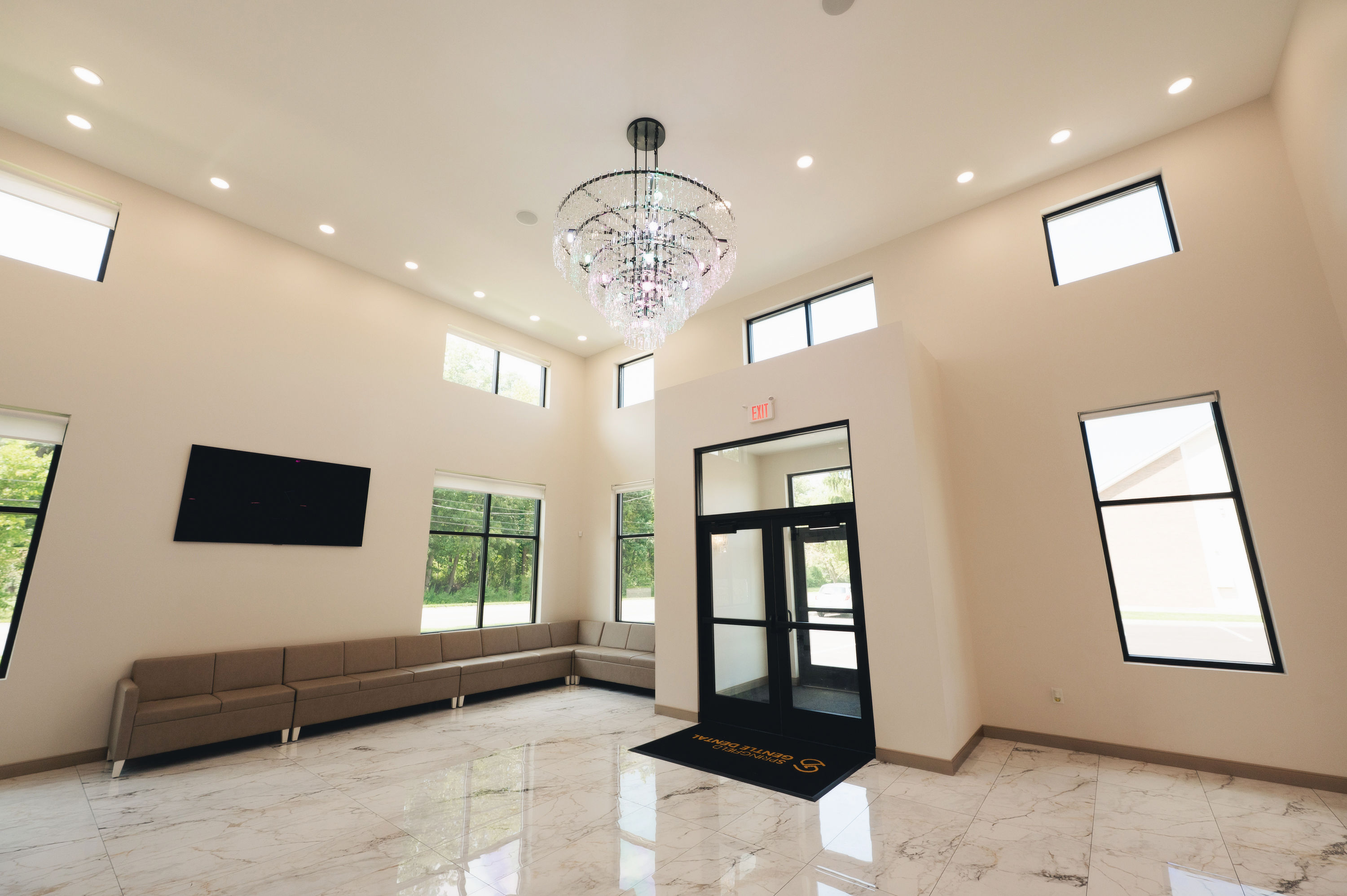 Bright spacious waiting area with beige sectional sofa, large windows, marble floor, and a crystal chandelier.