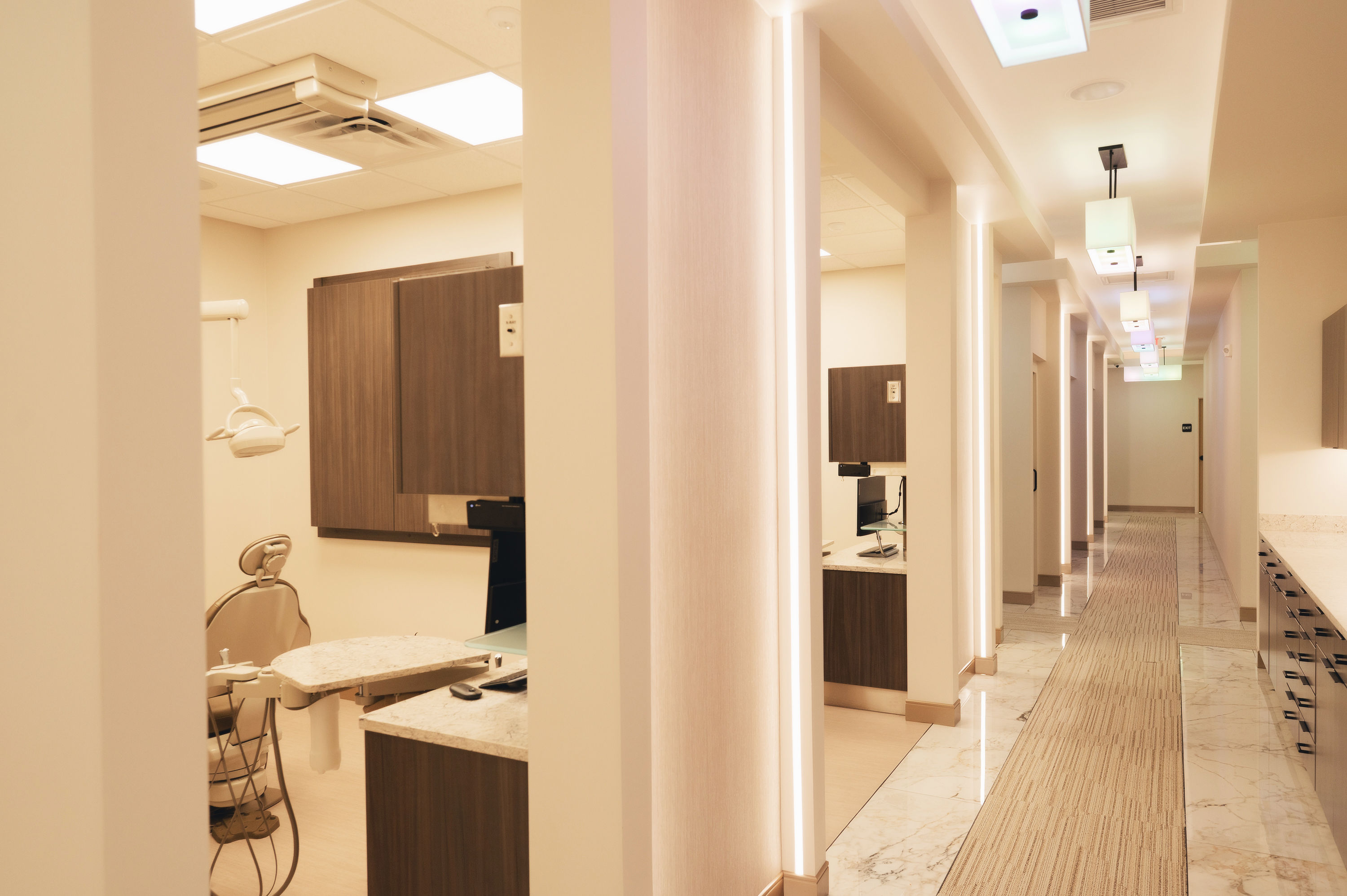 Modern dental office hallway with treatment rooms featuring dental chairs and equipment, soft lighting, and marble floors.
