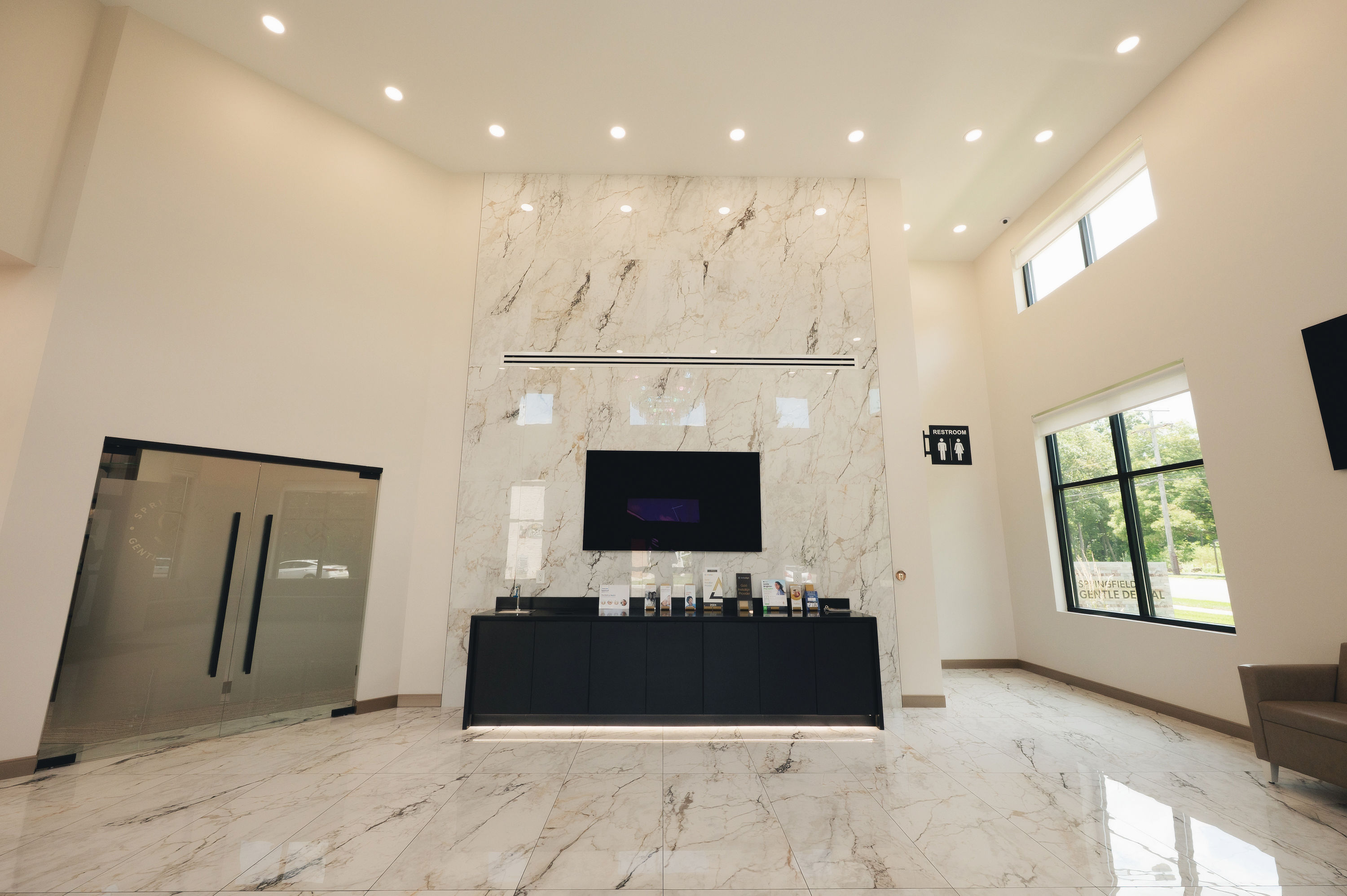 Modern dental office lobby with marble floors and walls, a black reception desk, large windows, and a flat-screen TV mounted above the desk.