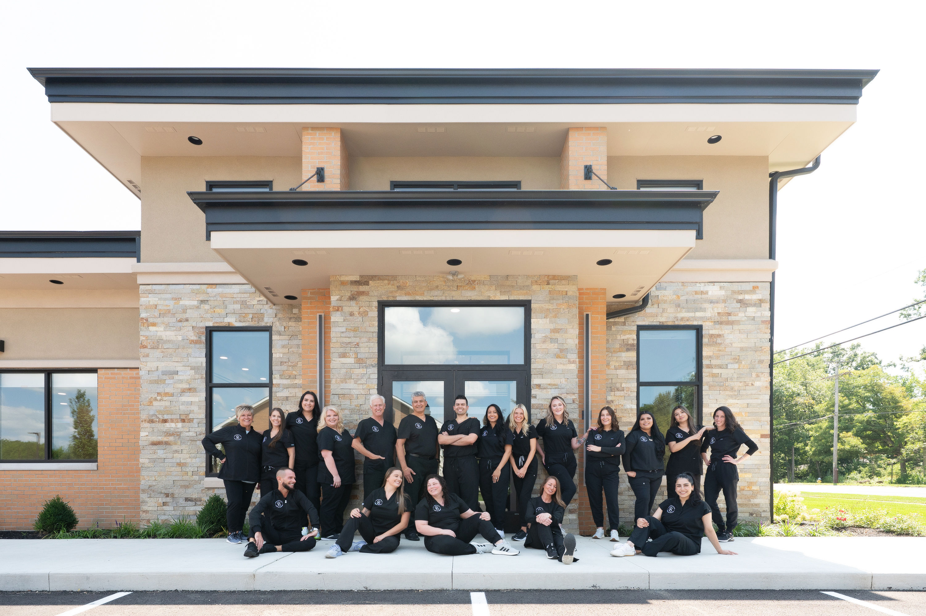 Group of smiling dental staff in black uniforms posing in front of a modern dental office building.