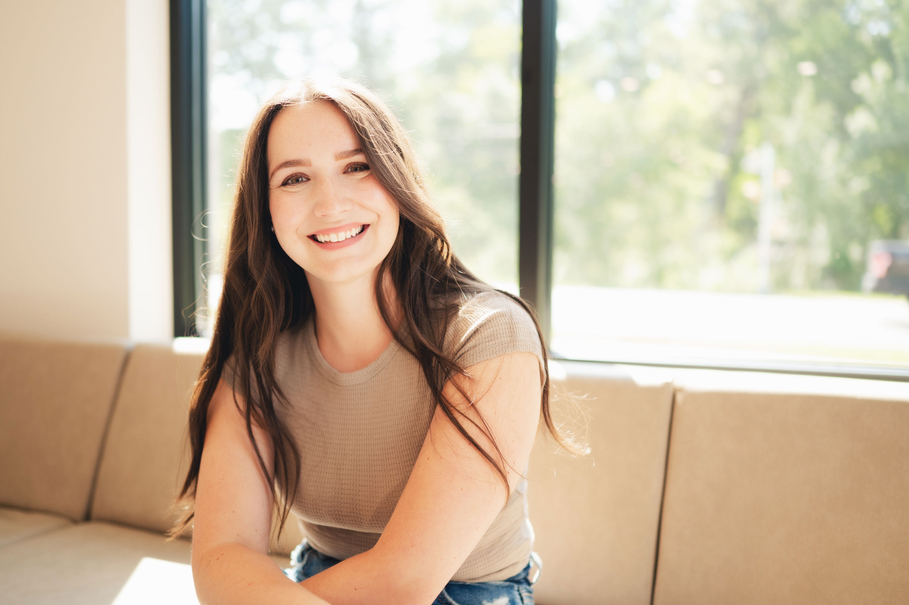 Smiling woman with long brown hair sitting on a beige couch in front of a large window with greenery outside.