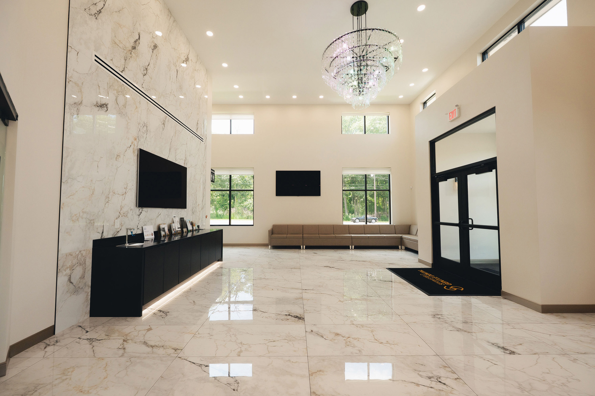 Bright modern lobby with marble floors, a chandelier, wall-mounted TVs, beige sectional sofa, and black double doors.