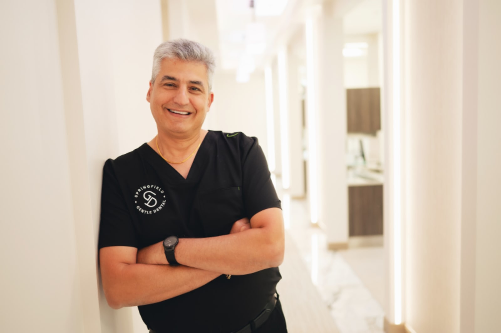 Male dentist in dark scrubs with practice logo, arms crossed, professional portrait