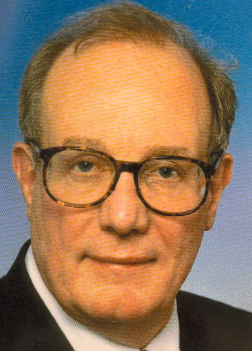 Philip Wood wearing large tortoiseshell glasses and a dark suit with white shirt against a blue background.