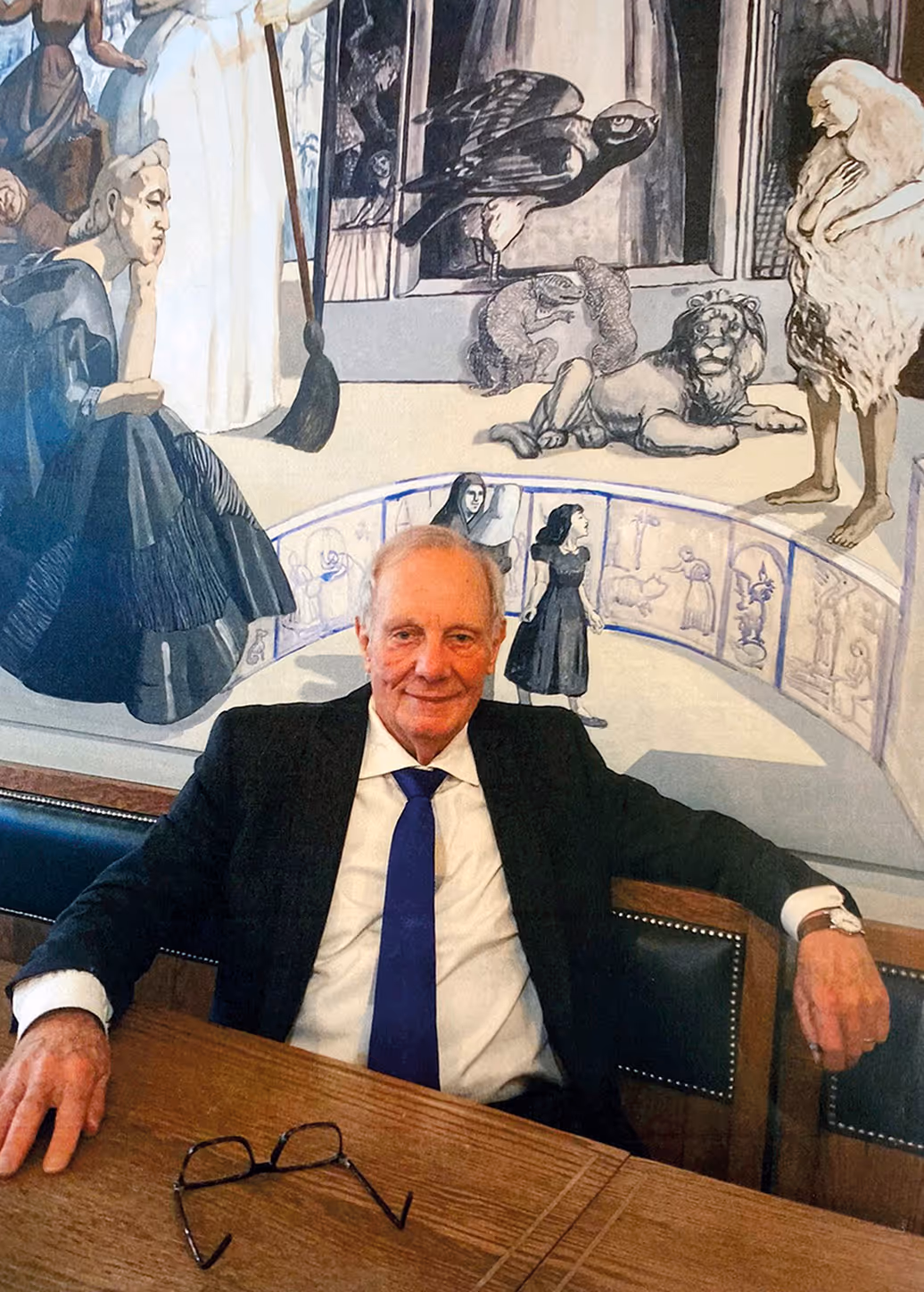 Elderly man in a suit with a blue tie sitting at a wooden table with eyeglasses on it, in front of a mural depicting various human and animal figures.