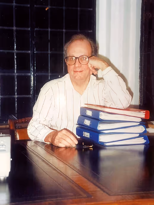 Philip Wood wearing glasses and a white striped shirt sitting at a dark wooden table with a stack of blue and red binders.