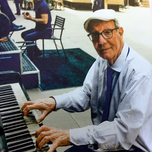 Elderly man wearing glasses, a white cap, and a white shirt playing an outdoor piano.
