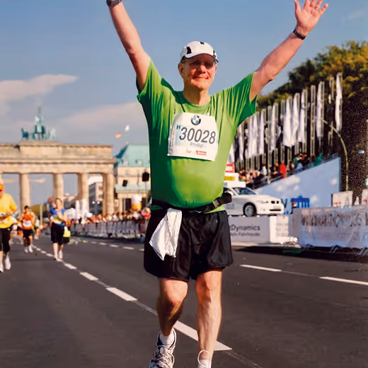A marathon runner wearing a green shirt and white cap raises his arms in celebration while running on a city street with spectators and Brandenburg Gate in the background.