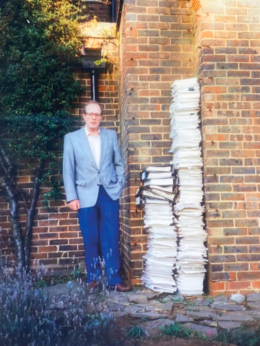 Man in a gray blazer standing outside next to two tall stacks of bundled paper against a brick wall.