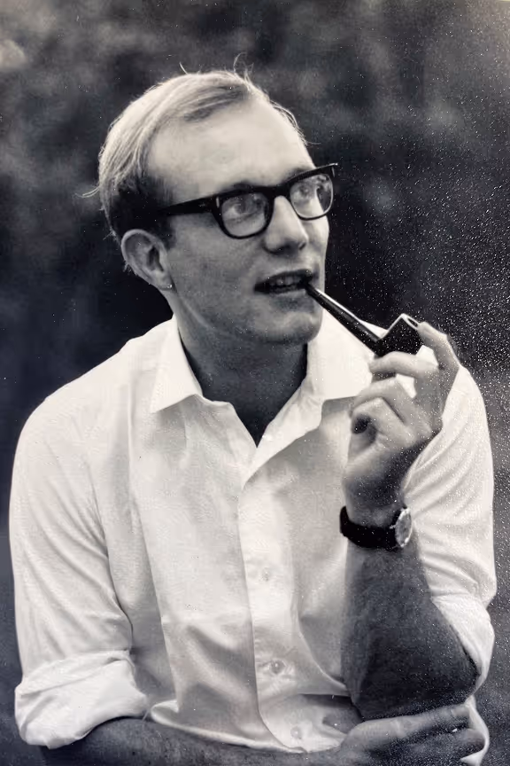 Black and white photo of a Philip Wood wearing glasses and a white shirt, holding a pipe near his mouth while looking to the side.