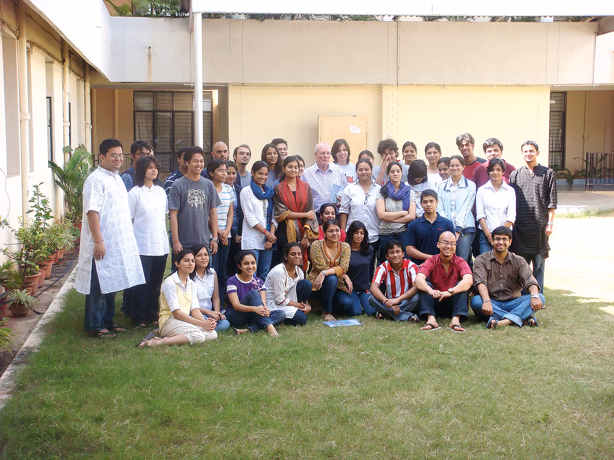 Group of diverse adults gathered outdoors on grass, smiling for a group photo in front of a building.