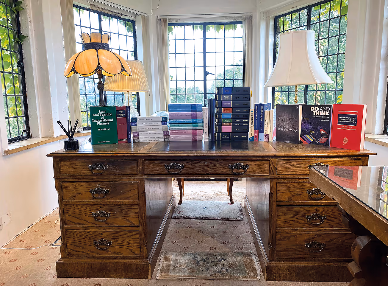 Antique wooden desk with stacks of legal books and two lamps in front of large leaded glass windows with visible greenery outside.