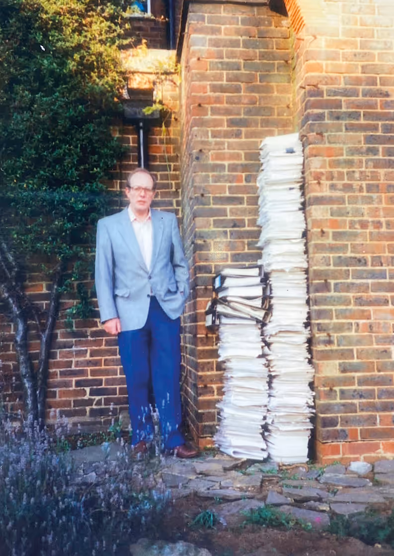 Philip Wood in a gray blazer and blue pants standing next to two tall stacks of papers and binders against a brick wall outside.